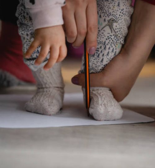 Checking baby foot size mother holding leg of her child on the blank paper drawing around with pencil to measure feet size family concept close up