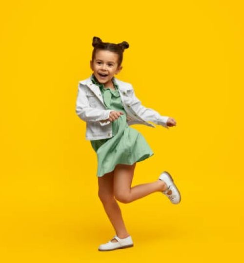Adorable little girl in trendy dress and jacket cheerfully smiling and twisting on one leg while dancing against bright yellow background